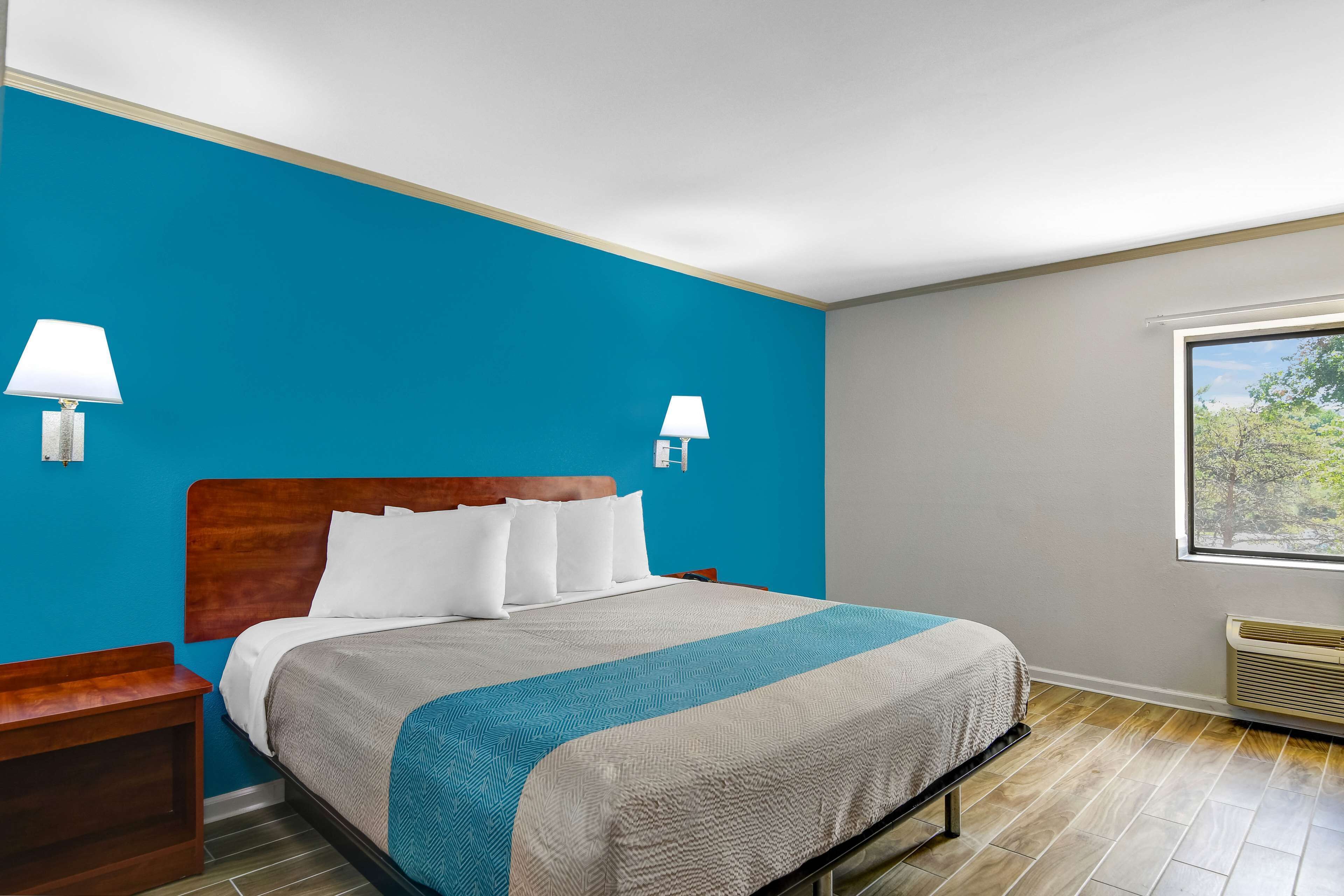 A hotel room with a bed, nightstand, two wall-mounted lamps, a window with a view, and a built-in heater.