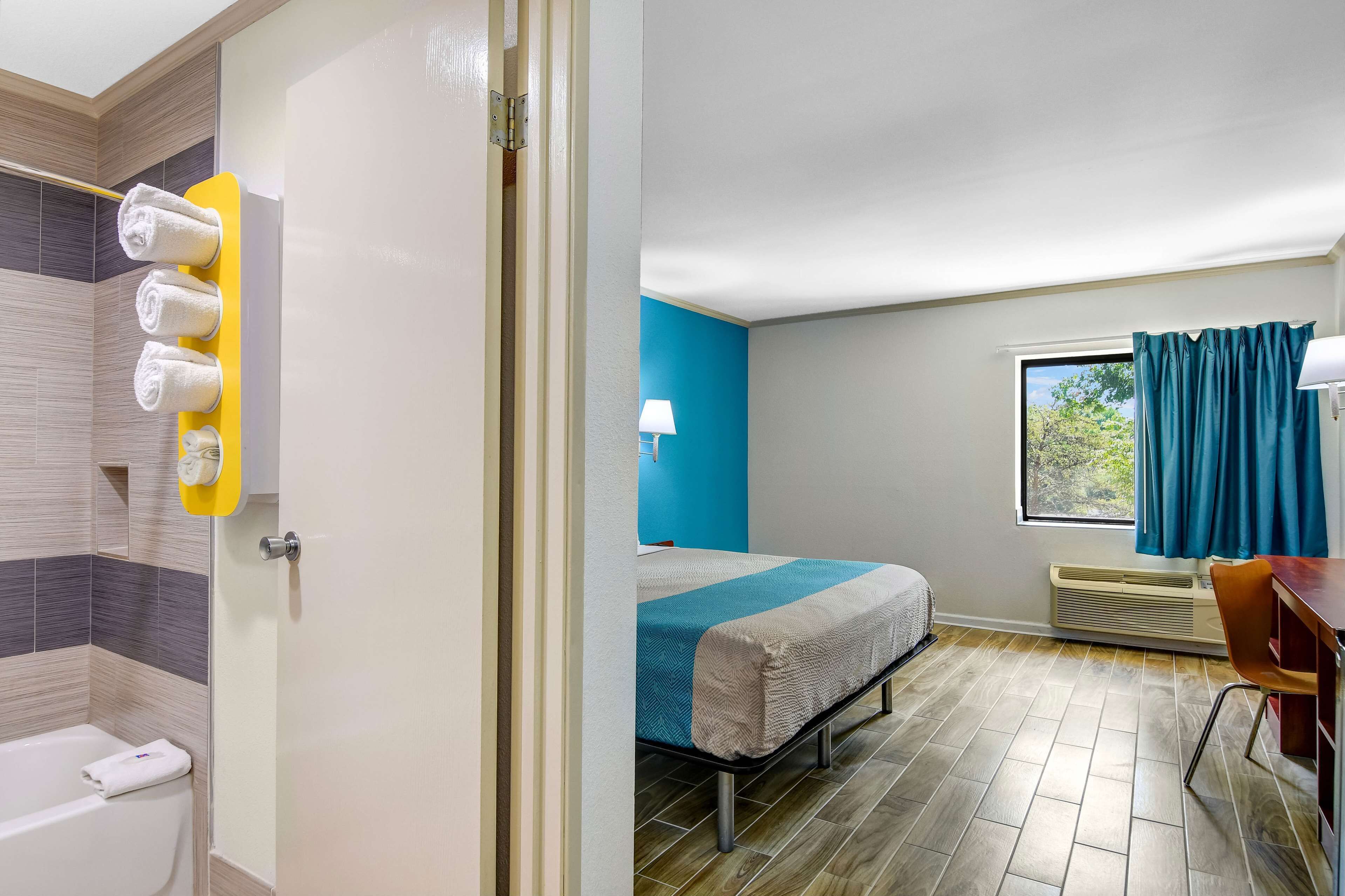 A hotel room with a bed, a desk, a chair, a window with curtains, a lamp, and a towel rack with towels.