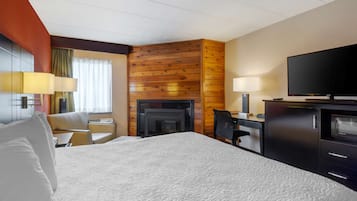 A hotel room with a large bed, a desk with a chair, a television, a fireplace, and a desk lamp.
