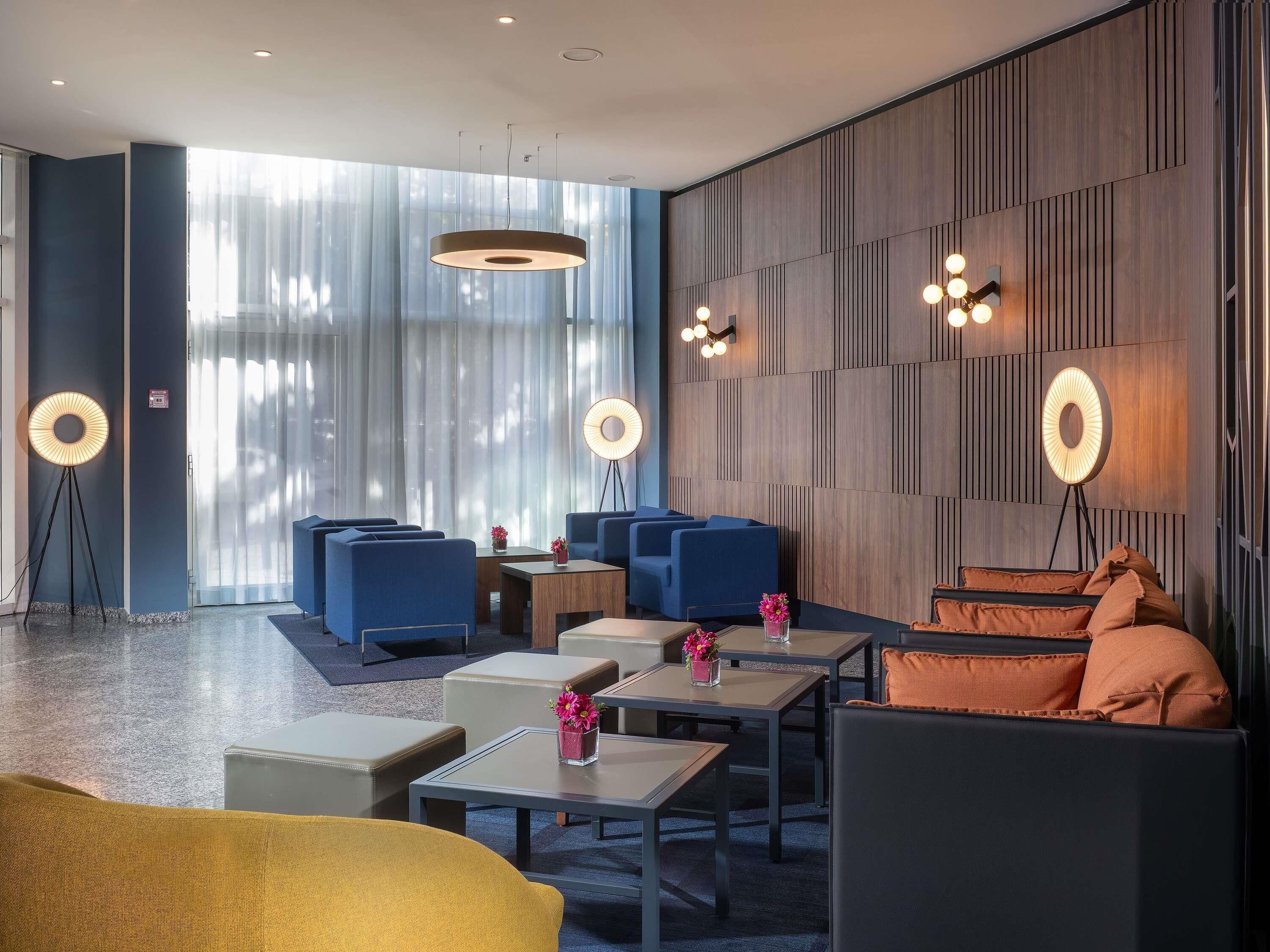 A modern hotel lobby with seating areas, a reception desk, and a large window with curtains.