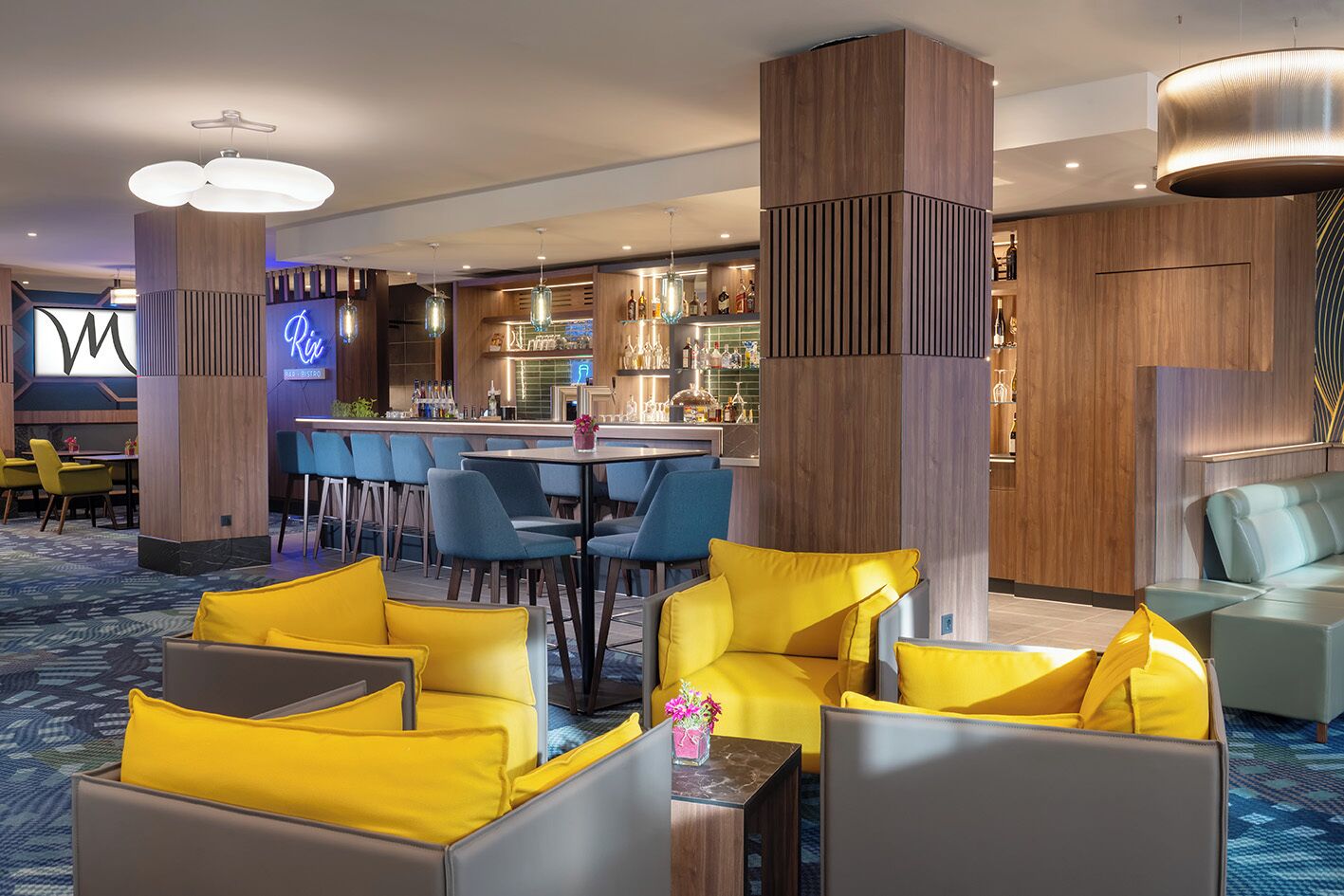 A modern hotel lobby with a bar area, seating with yellow cushions, and a neon sign.