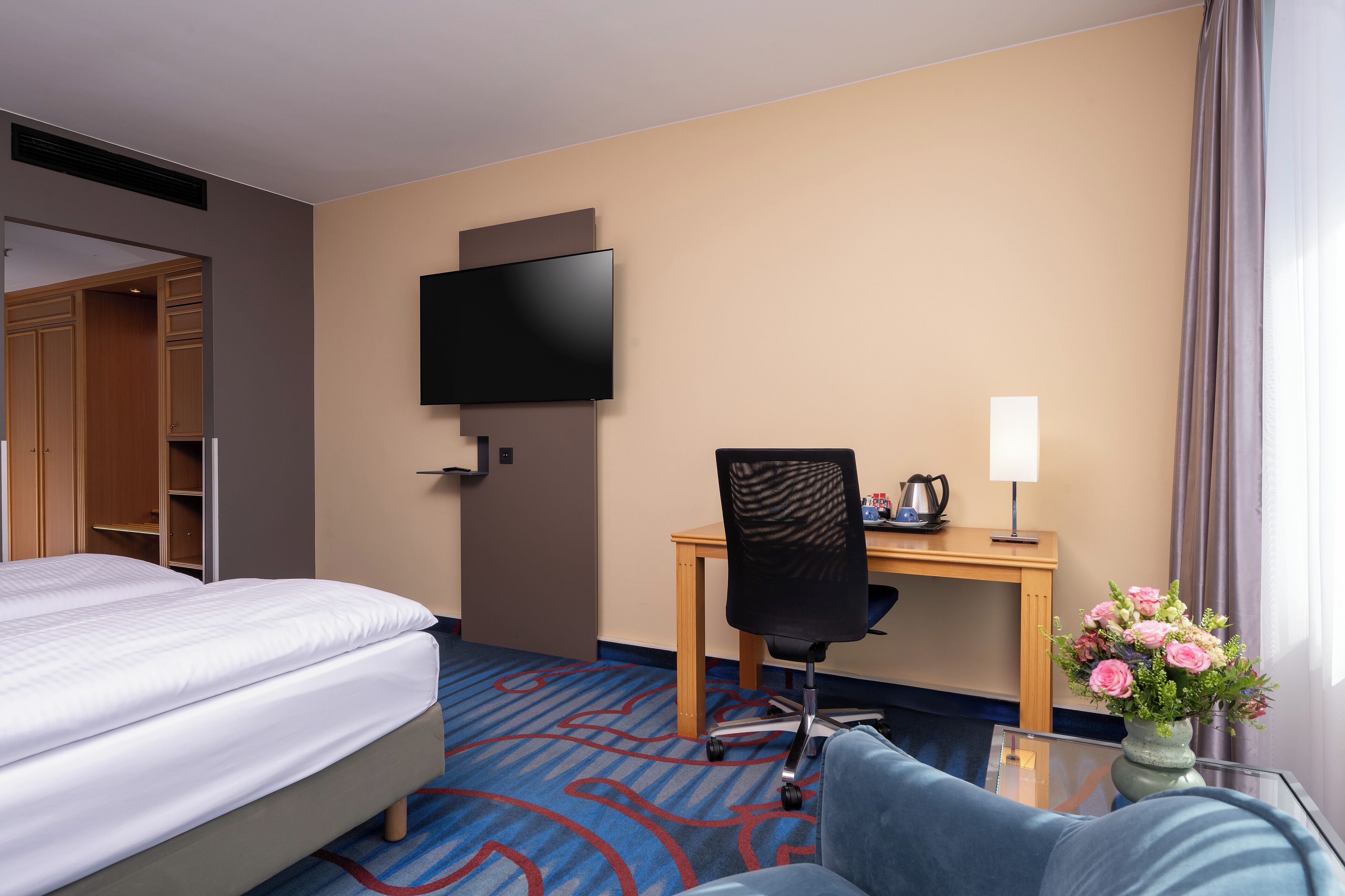 Una habitación de hotel con cama, escritorio con silla, televisor de pantalla plana, lámpara y un jarrón con flores.