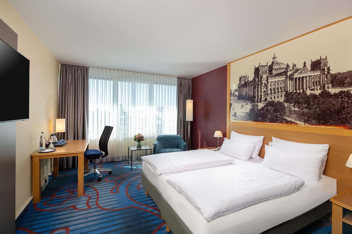 Hotelkamer met een groot bed, een bureau, een stoel, een klein tafeltje met een vaas, en een groot raam met gordijnen.