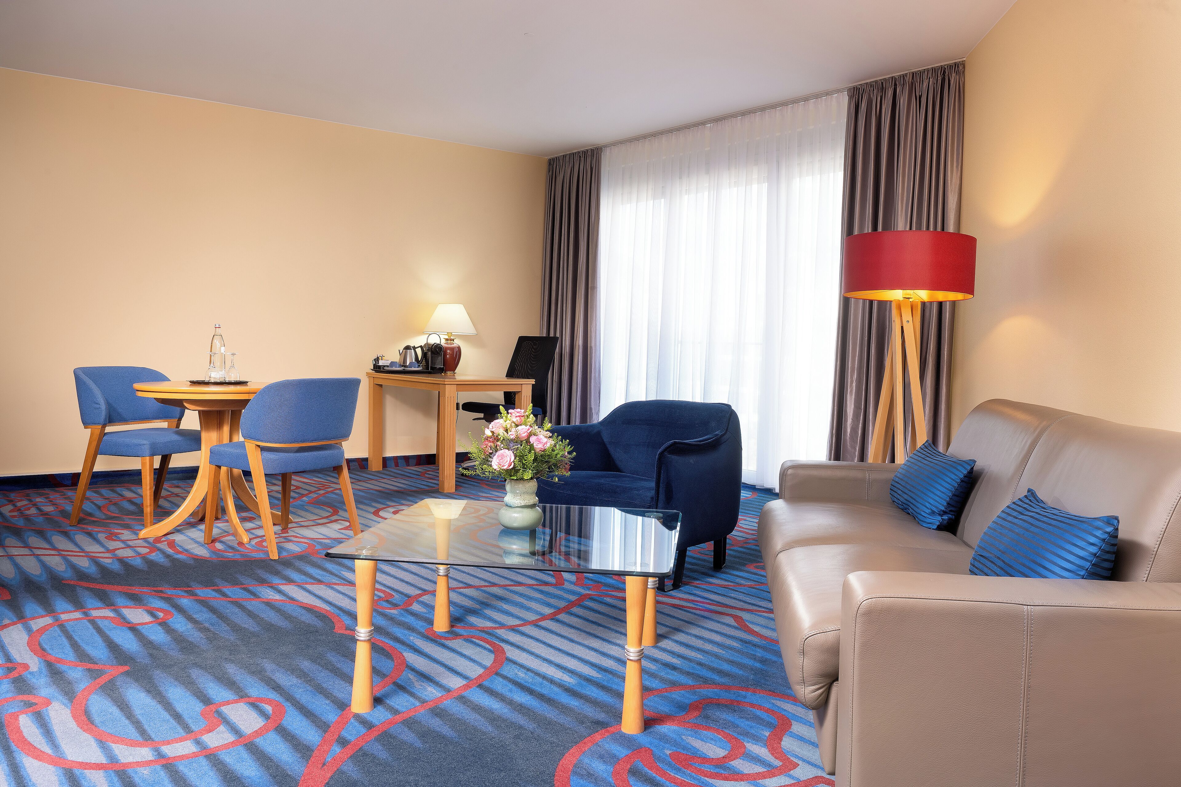 Una habitación de hotel con un sofá color beige, un sillón azul, una mesa de centro de vidrio, un escritorio pequeño con una l ámpara y un ventanal con cortinas.