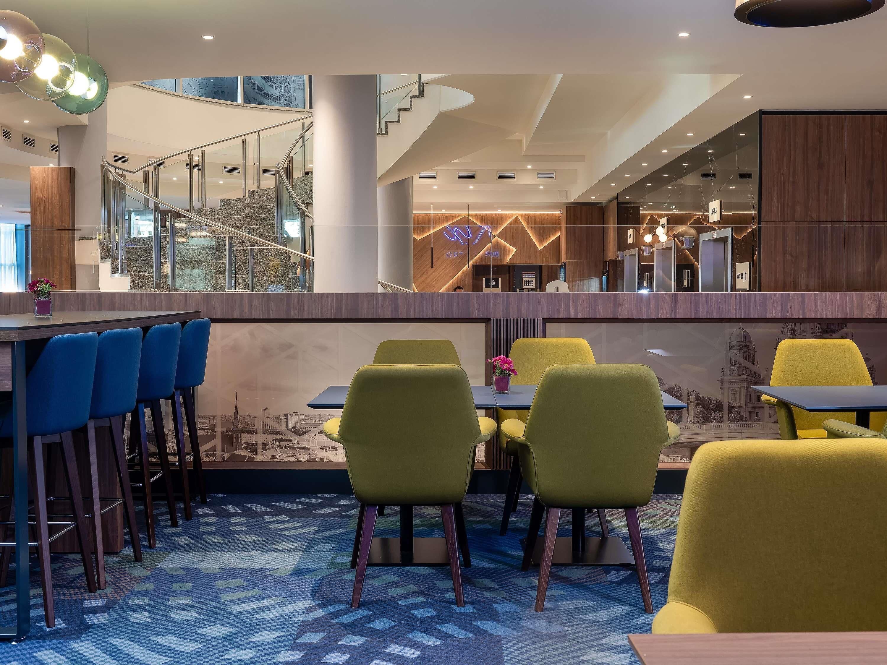 A modern hotel lobby with a high ceiling, a staircase, and a seating area with yellow chairs and a patterned carpet.