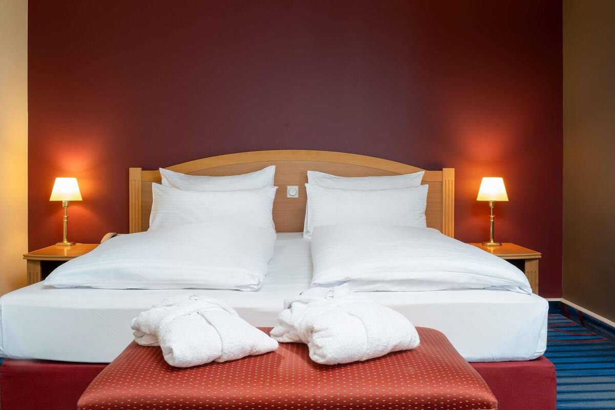 Een tweepersoonsbed met witte lakens, twee bedlampjes en een rode poef met handdoeken.