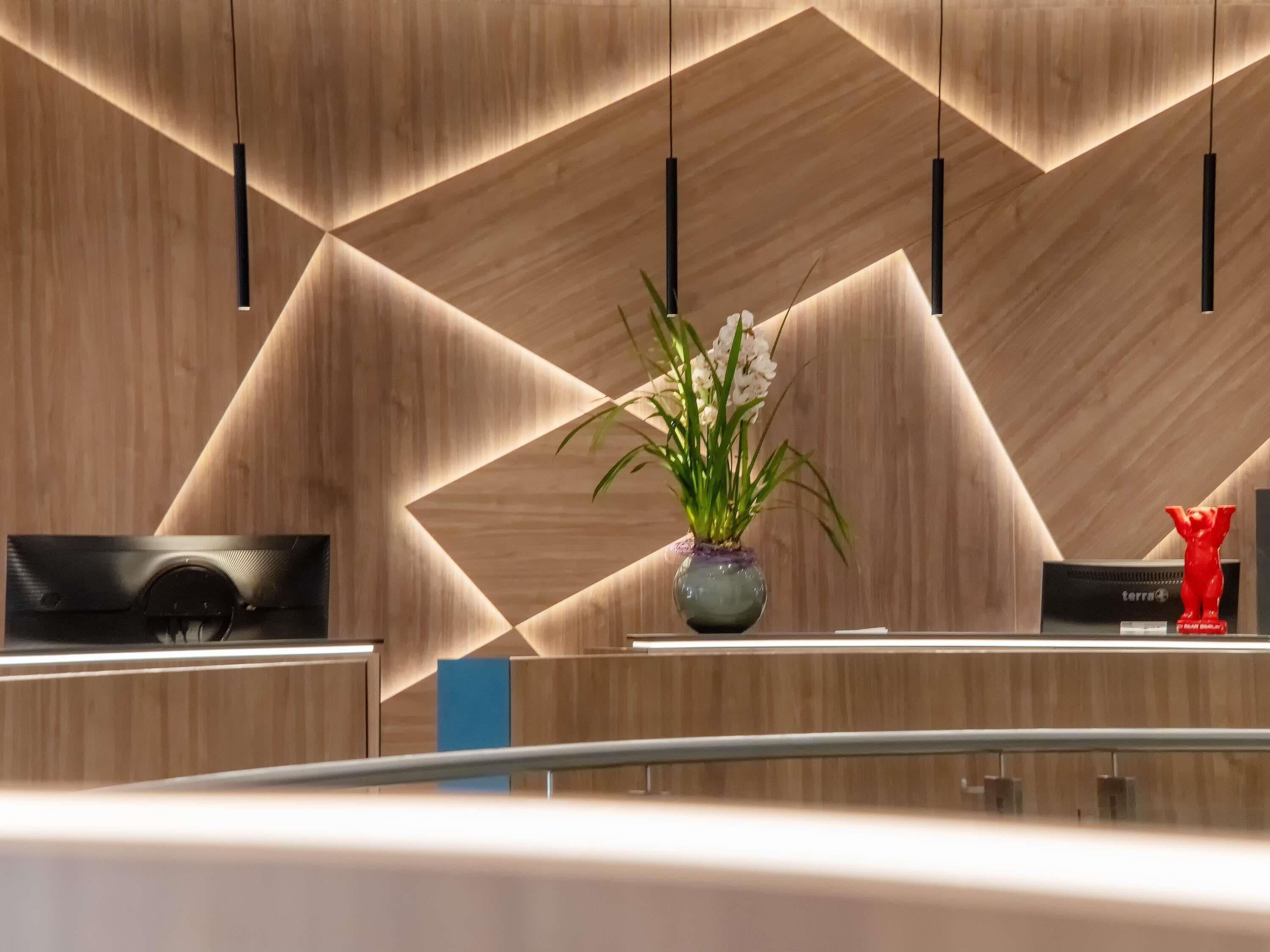 Un mostrador de recepción moderno con un diseño geométrico de pared de madera, una planta en maceta y una estatua roja.