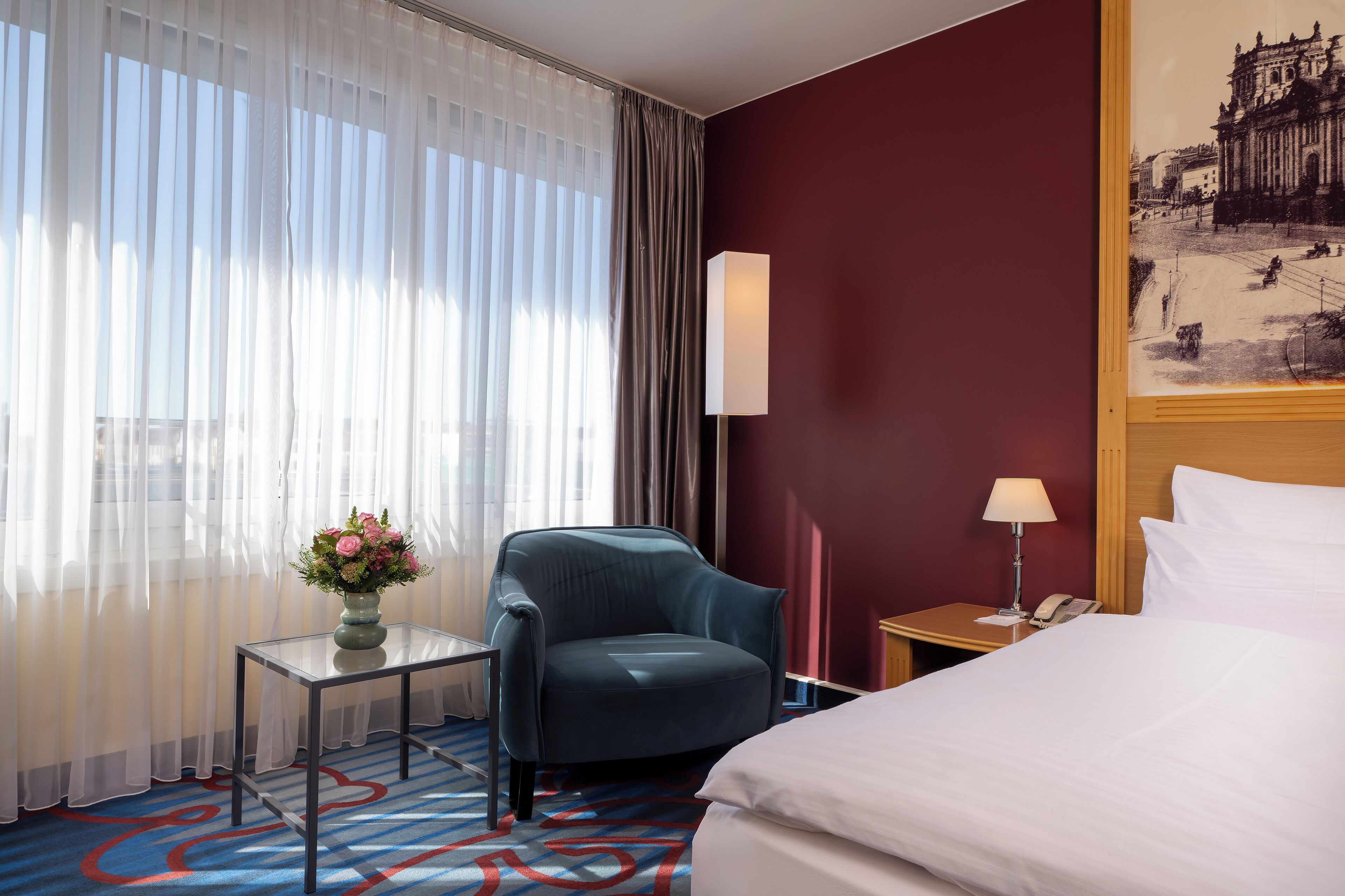 A hotel room with a bed, a desk with a lamp, a chair, a vase with flowers, and a painting on the wall.