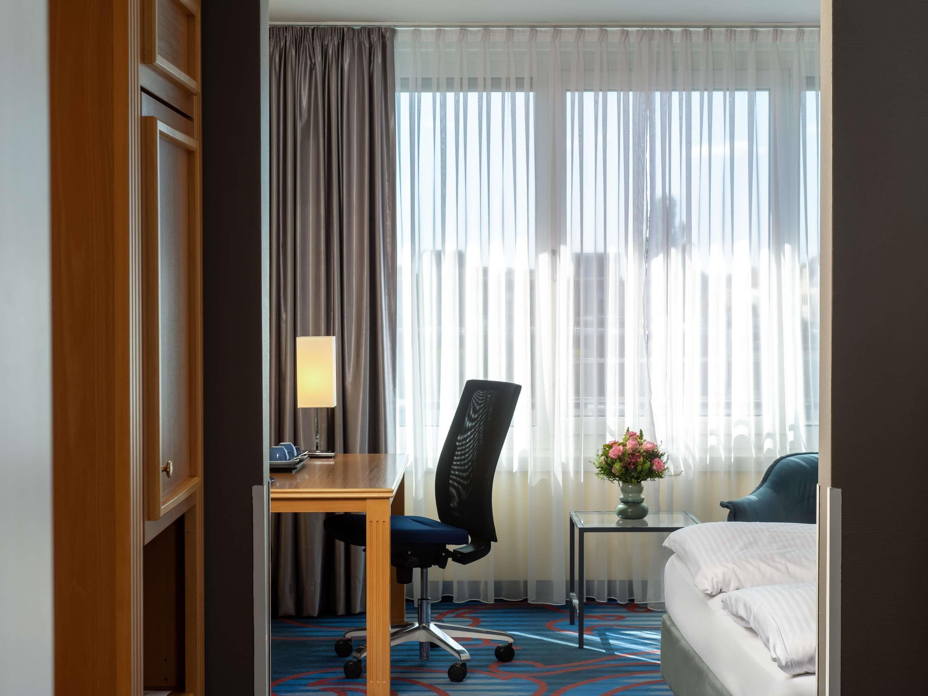 Ein Hotelzimmer mit einem Bett, einem Schreibtisch mit Stuhl, einer Lampe, einer Vase mit Blumen und einem Fenster mit Vorhängen.
