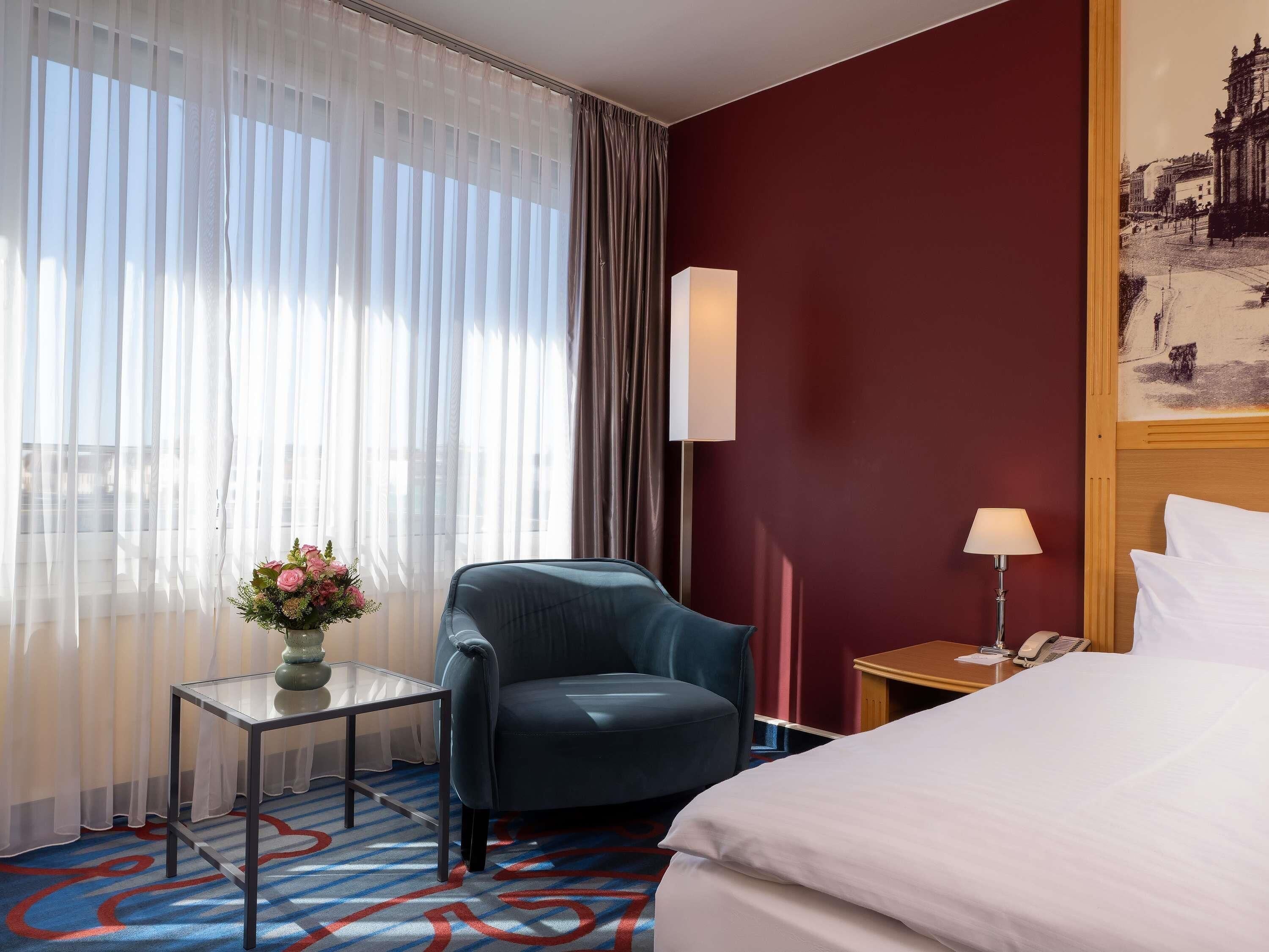 A hotel room with a bed, a chair, a small table with a vase of flowers, a lamp, and a painting on the wall.