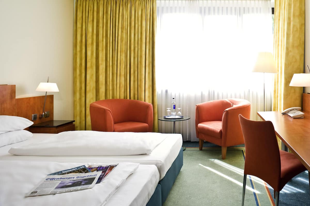 Habitación de hotel con dos camas, un escritorio, dos sillas, una lámpara y un ventanal con cortinas.