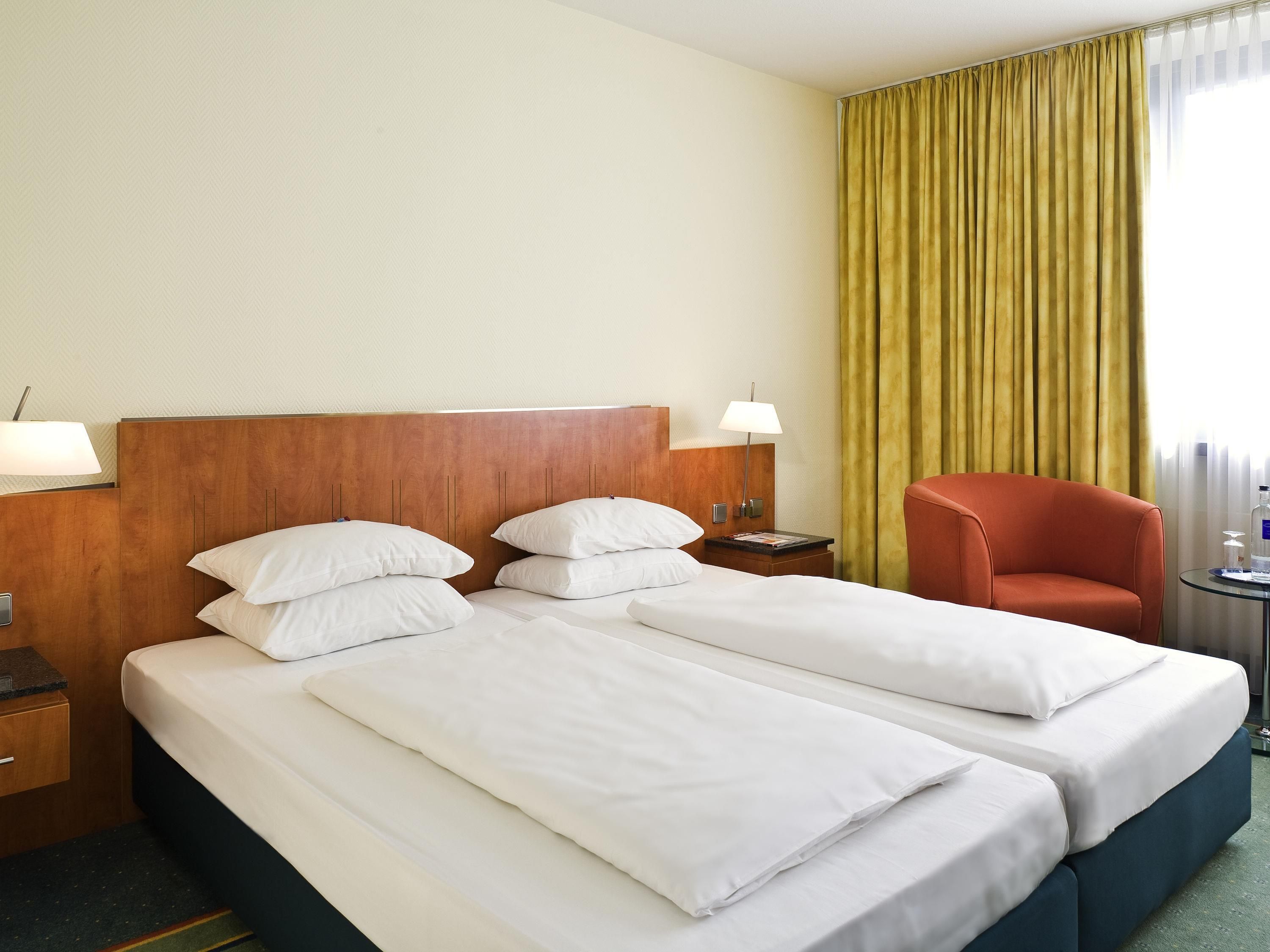 A hotel room with two beds, a wooden headboard, a nightstand with a lamp, a chair, and a window with curtains.