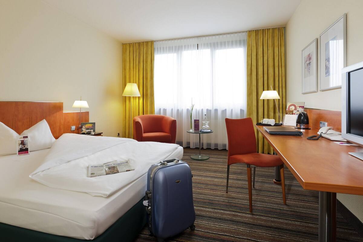 Habitación de hotel con cama, escritorio, dos sillas, lámpara de escritorio, televisor y ventana con cortinas.
