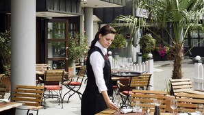 An outdoor dining area with wooden tables and chairs, a waiter attending to a table, and potted plants around. - Mercure Hotel Offenburg am Messeplatz (Offenburg)