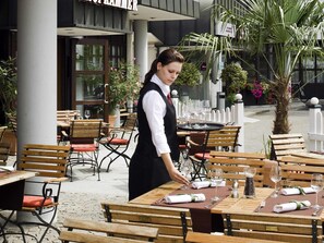 An outdoor dining area with wooden tables and chairs, a waiter attending to a table, and potted plants around. - Mercure Hotel Offenburg am Messeplatz (Offenburg)