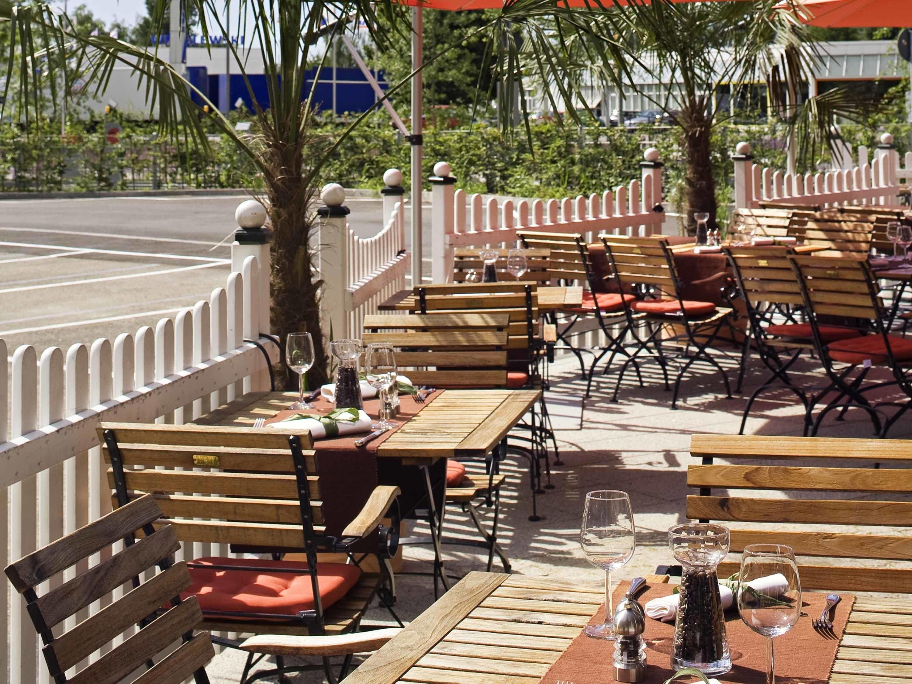 Een buitenterras met houten tafels en stoelen, gerangschikt in rijen, omgeven door een witte schutting.