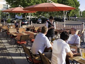 Sitzgelegenheiten im Freien mit Tischen und Stühlen, Sonnenschirmen und einem weißen Zaun im Hintergrund.