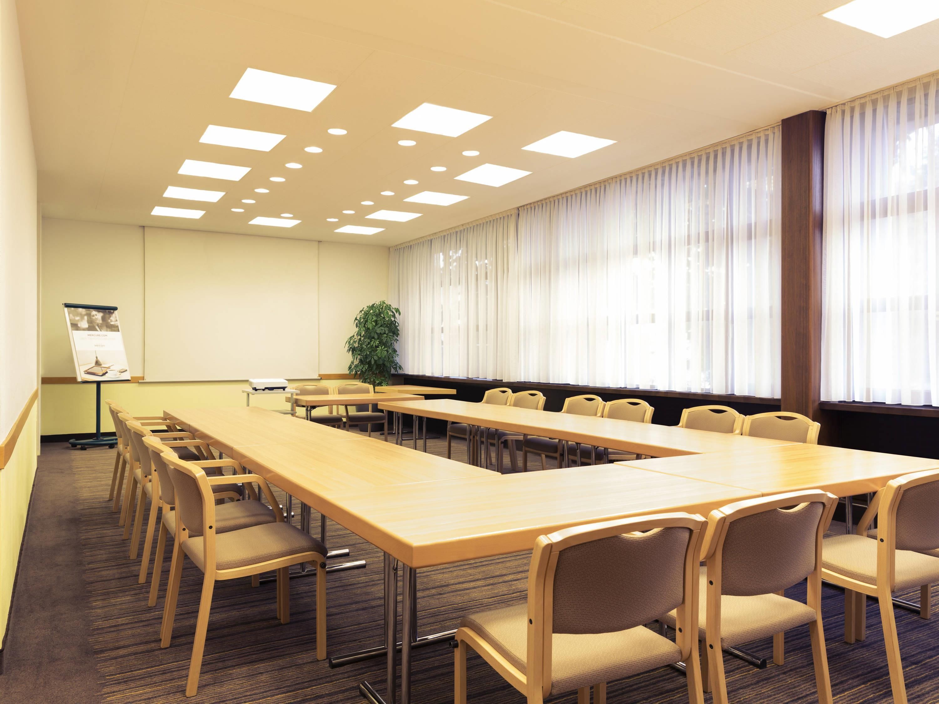 Ein Konferenzraum mit langem Tisch, Stühlen, einer Projektionsleinwand und einer Pflanze.