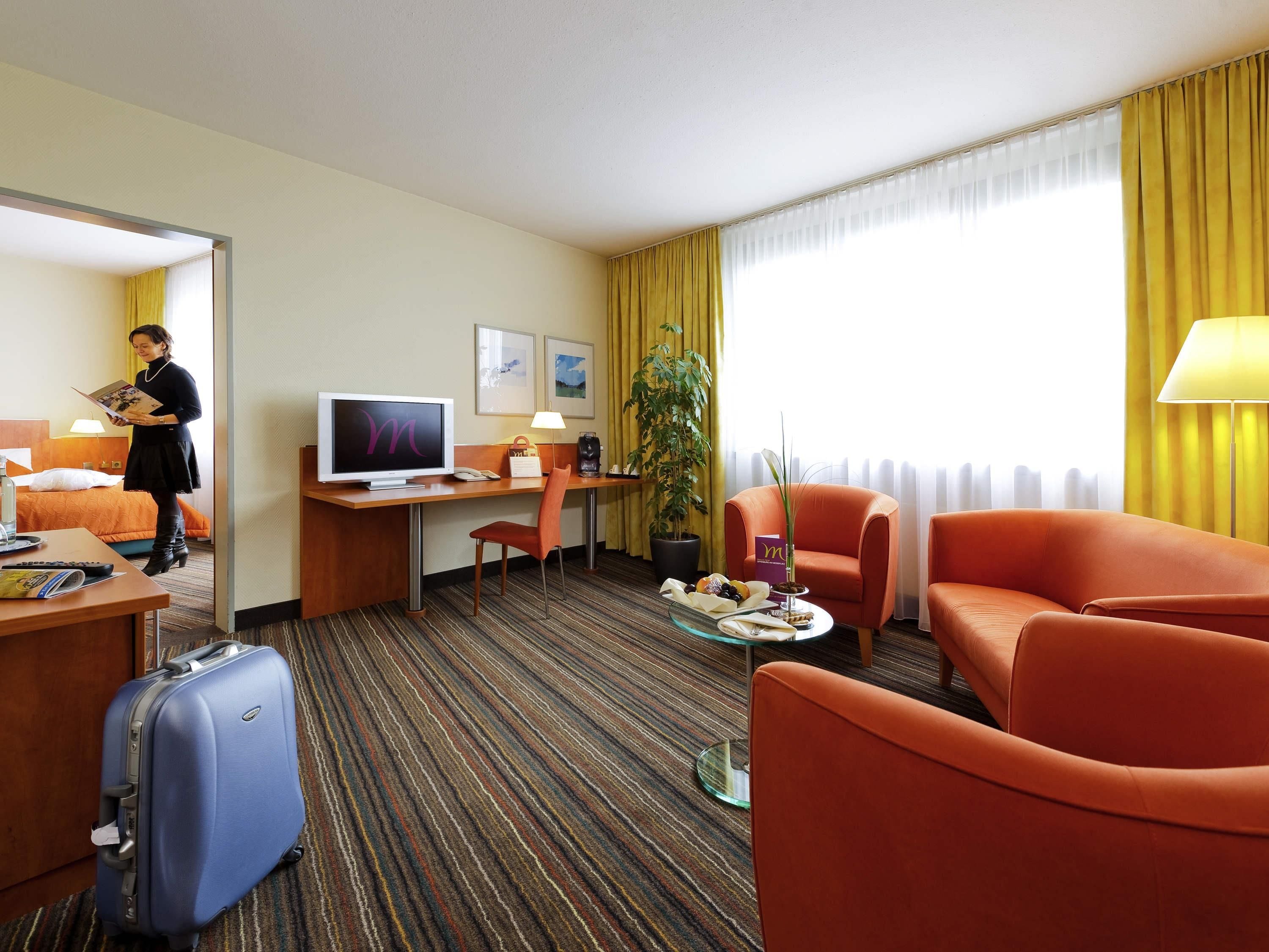Ein Hotelzimmer mit Bett, Schreibtisch, Fernseher, Sessel und Koffer.