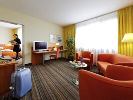 Habitación de hotel con cama, escritorio, televisión, sillón y maleta.