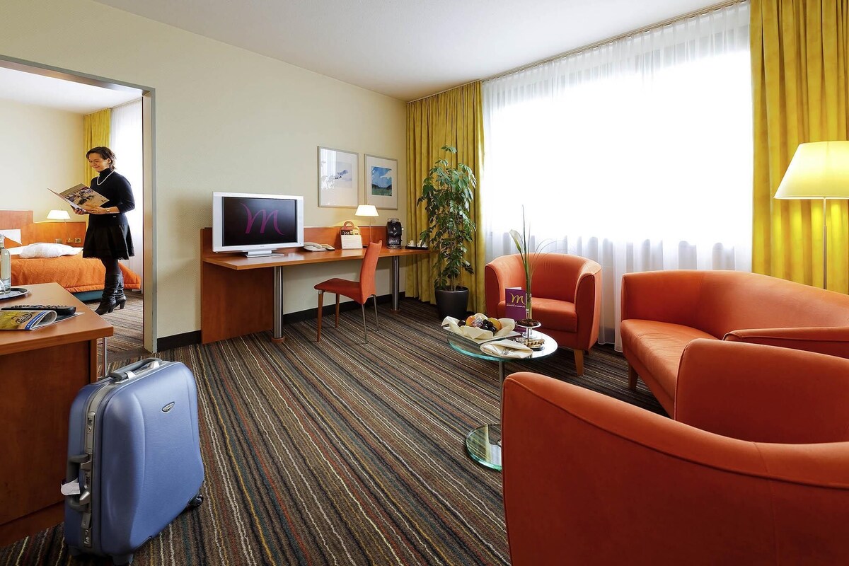 A hotel room with a bed, desk, television, two chairs, a suitcase, and a large window with curtains.