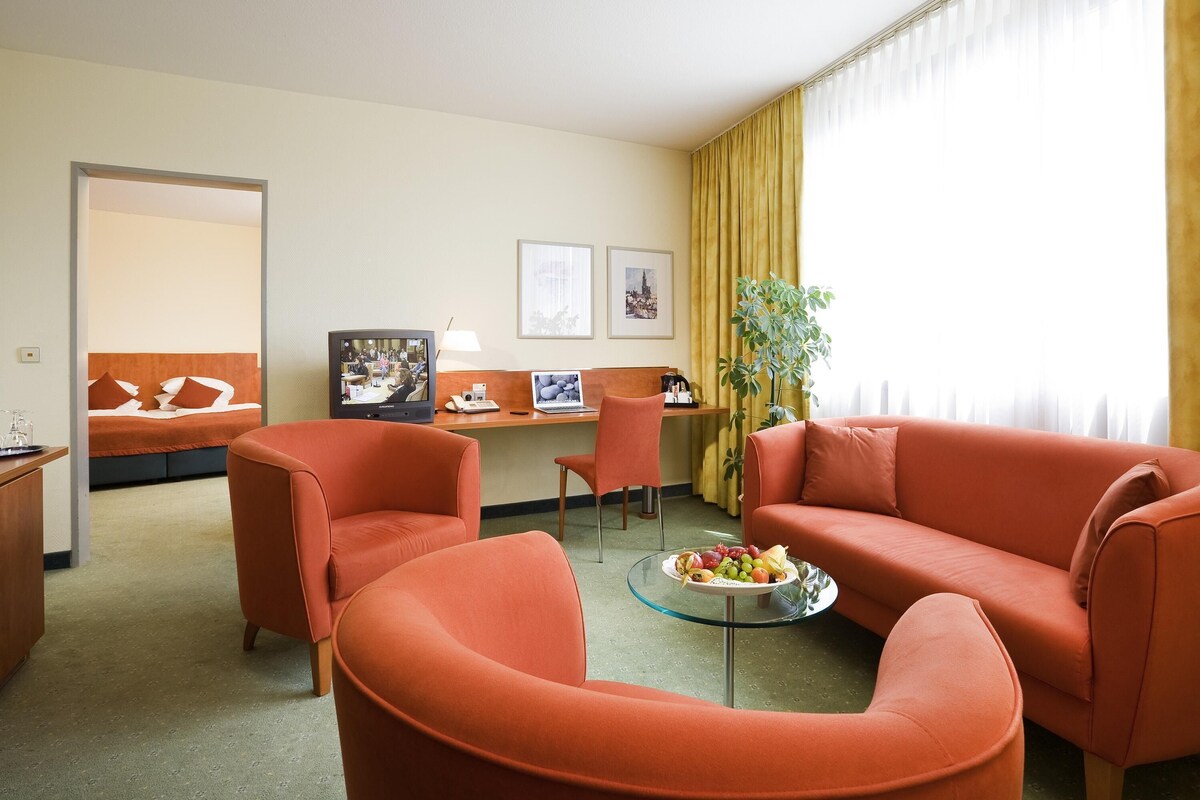 A hotel room with a bed, a desk with a computer, a television, two orange armchairs, a glass coffee table with fruit, and a large window with curtains.