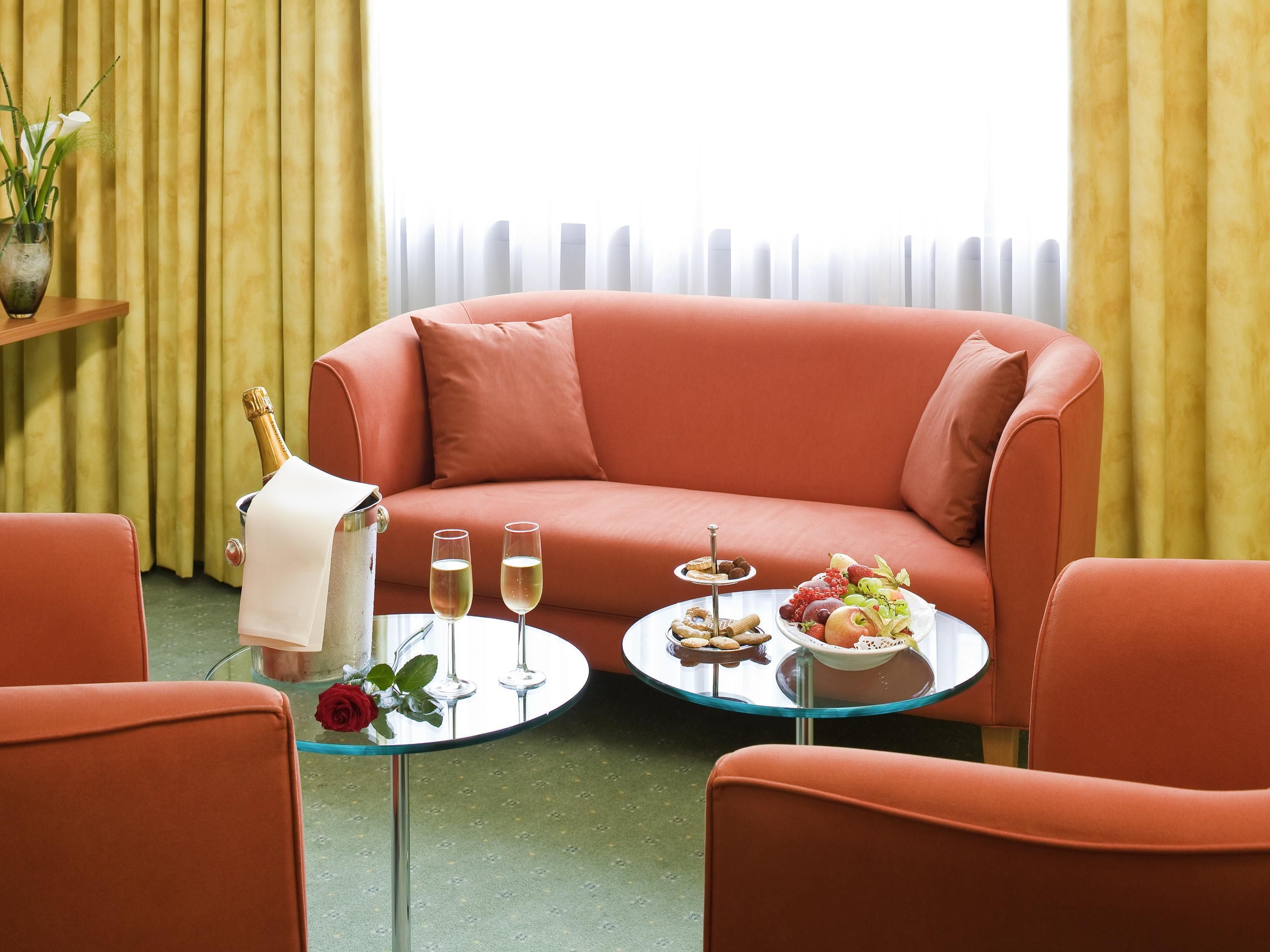 Una habitación con un sofá rojo, una mesa de vidrio con una bandeja de frutas, dos copas de champán y un jarrón con una rosa.