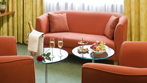 A room with a red sofa, a glass table with a fruit platter, two glasses of champagne, and a vase with a rose. - Mercure Hotel Offenburg am Messeplatz (Offenburg)