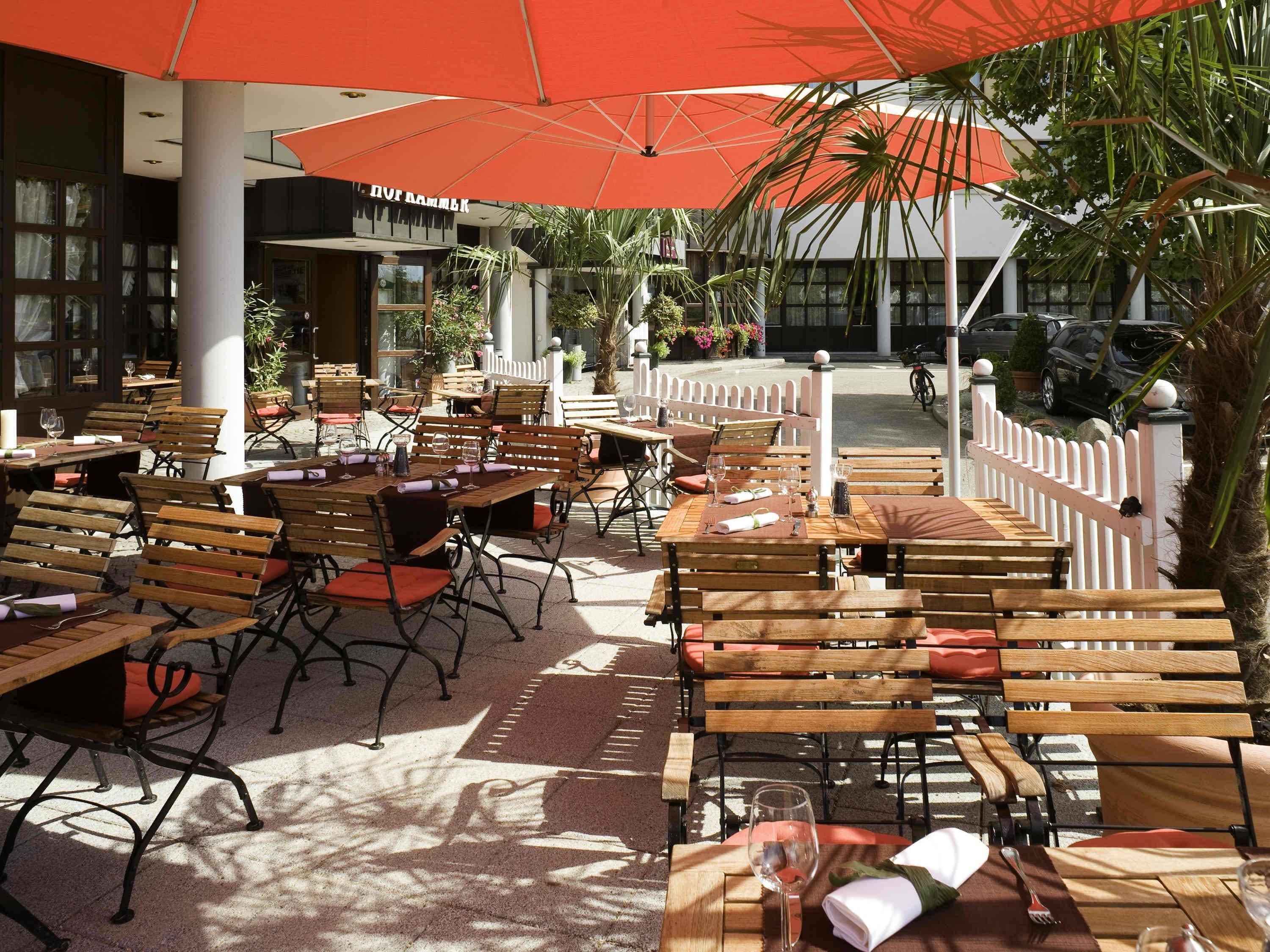 An outdoor dining area with wooden tables and chairs, umbrellas, and a white fence.