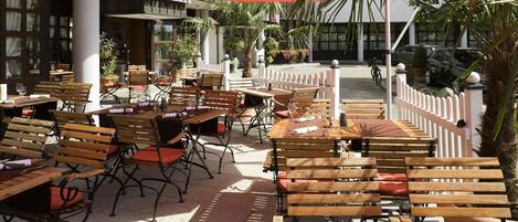 An outdoor dining area with wooden tables and chairs, umbrellas, and a white fence.