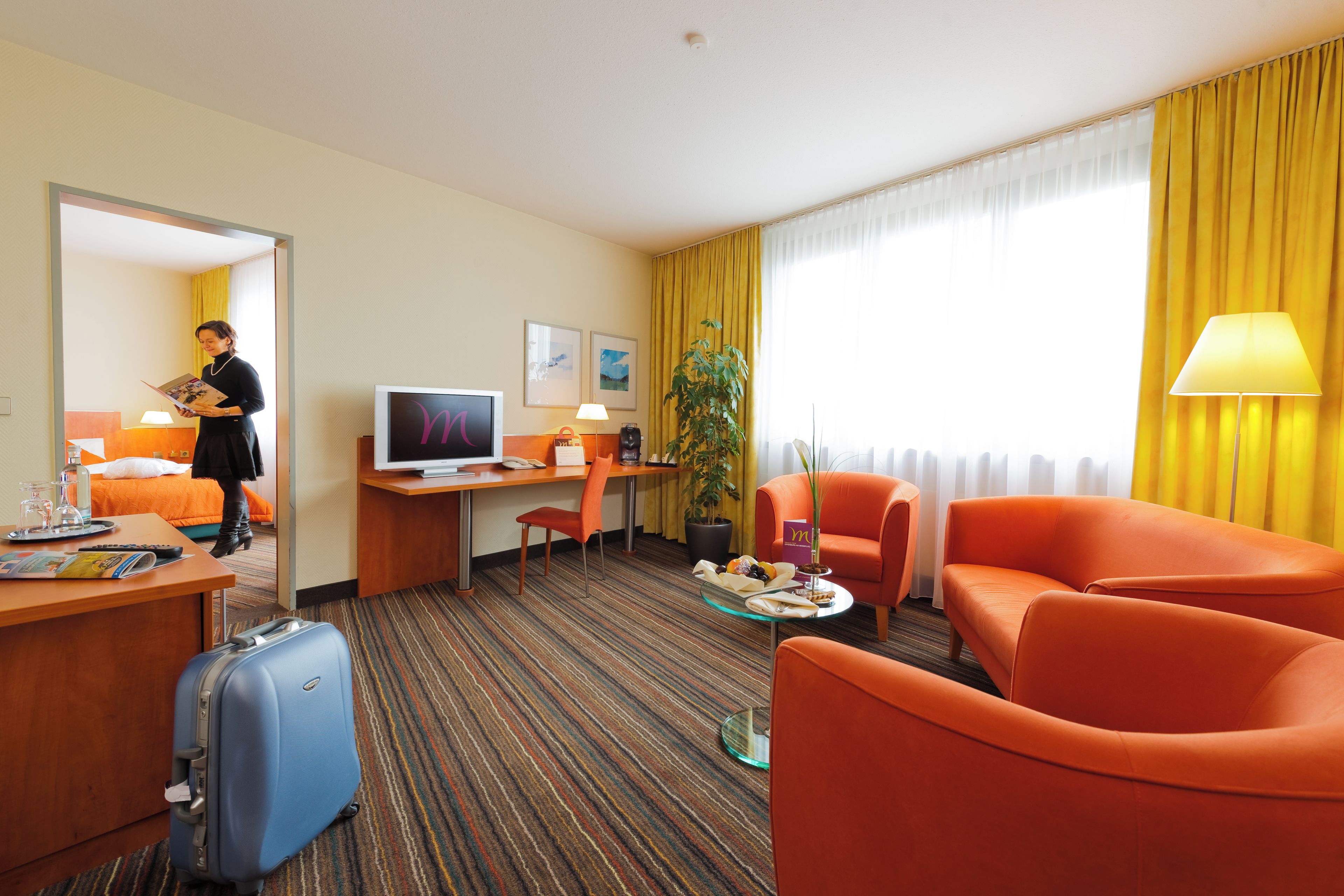 Habitación de hotel con cama, escritorio con computadora, televisor, zona de estar con sillas naranjas, mesita con refrigerios, planta en maceta y maleta.