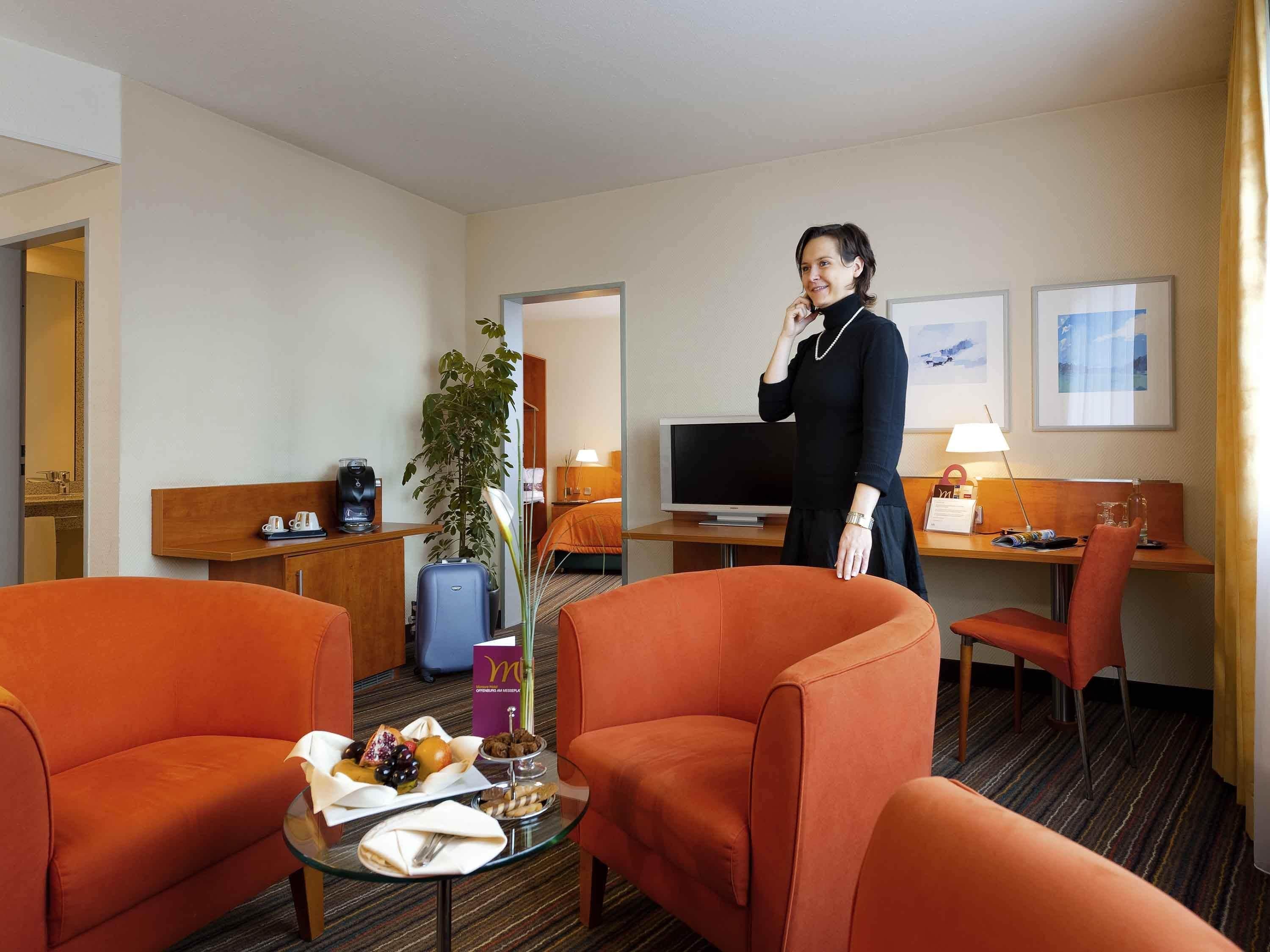 A hotel room with orange armchairs, a small table with breakfast, a TV, and a desk with a lamp.