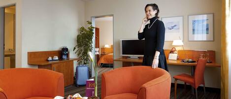 Habitación de hotel con sillones naranjas, una mesita con desayuno, un televisor y un escritorio con lámpara.