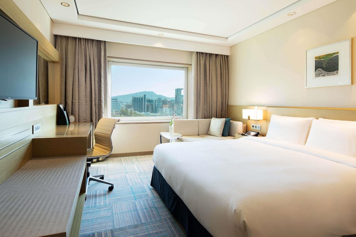 A hotel room with a bed, a desk, a chair, a TV, a window with curtains, a lamp, and a painting on the wall.
