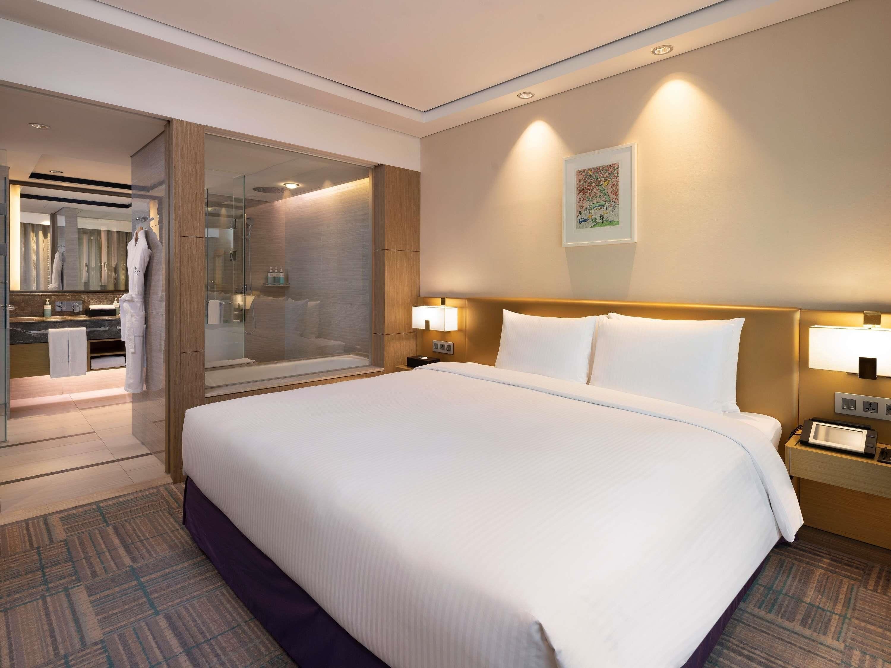 A hotel room with a large bed, bedside lamps, a nightstand, a glass shower enclosure, a bathroom with a mirror, and a framed picture on the wall.