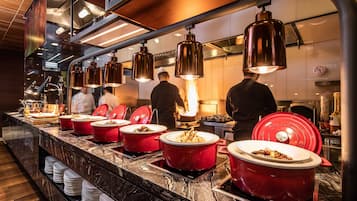 Un buffet disposé sur un comptoir en marbre, avec plusieurs plats de service rouges à réchaud, un éclairage industriel suspendu au plafond et une cuisine visible en arrière-plan.