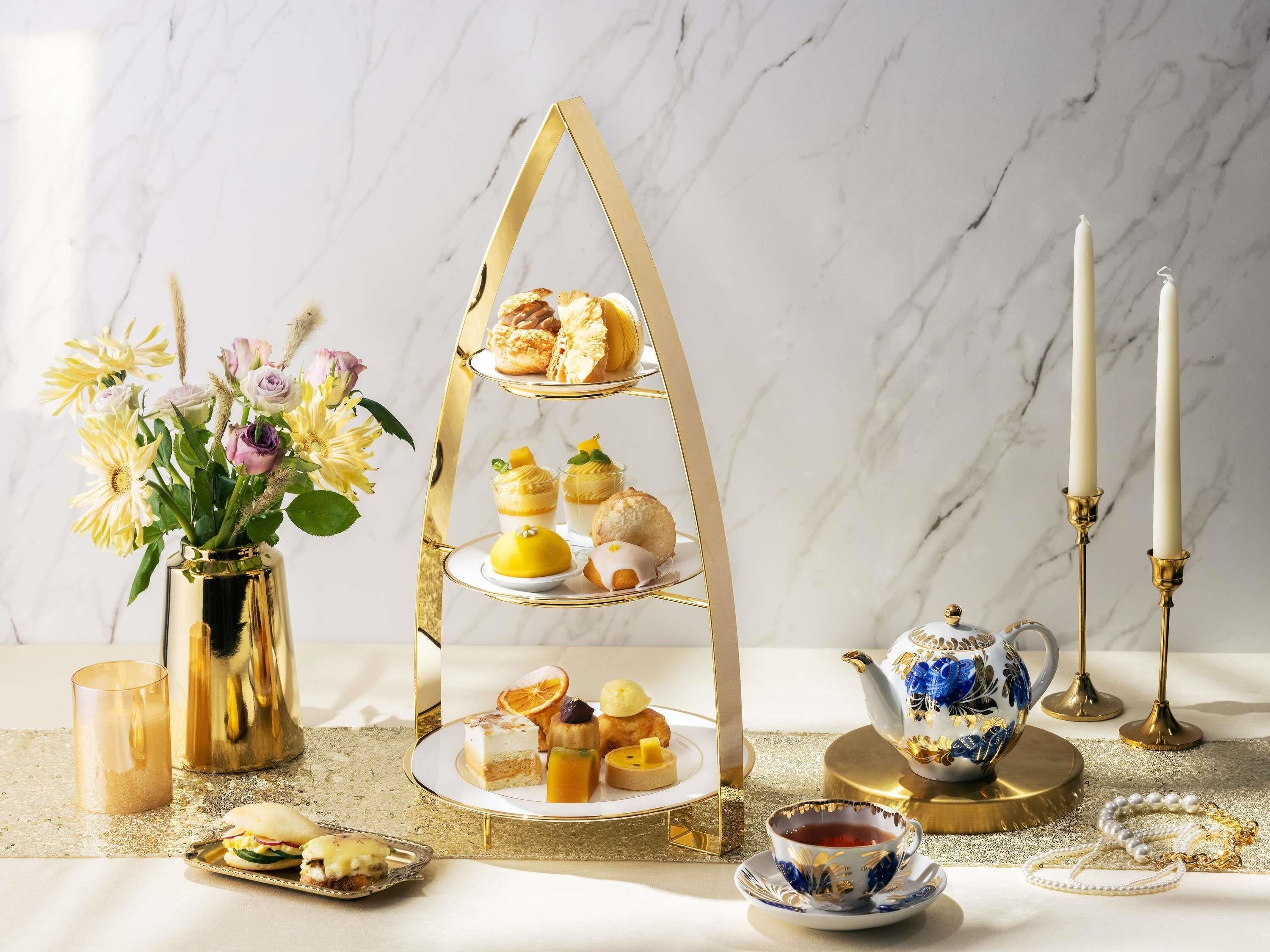 A three-tiered dessert stand with pastries, a vase of flowers, a teapot, and candles on a marble table.