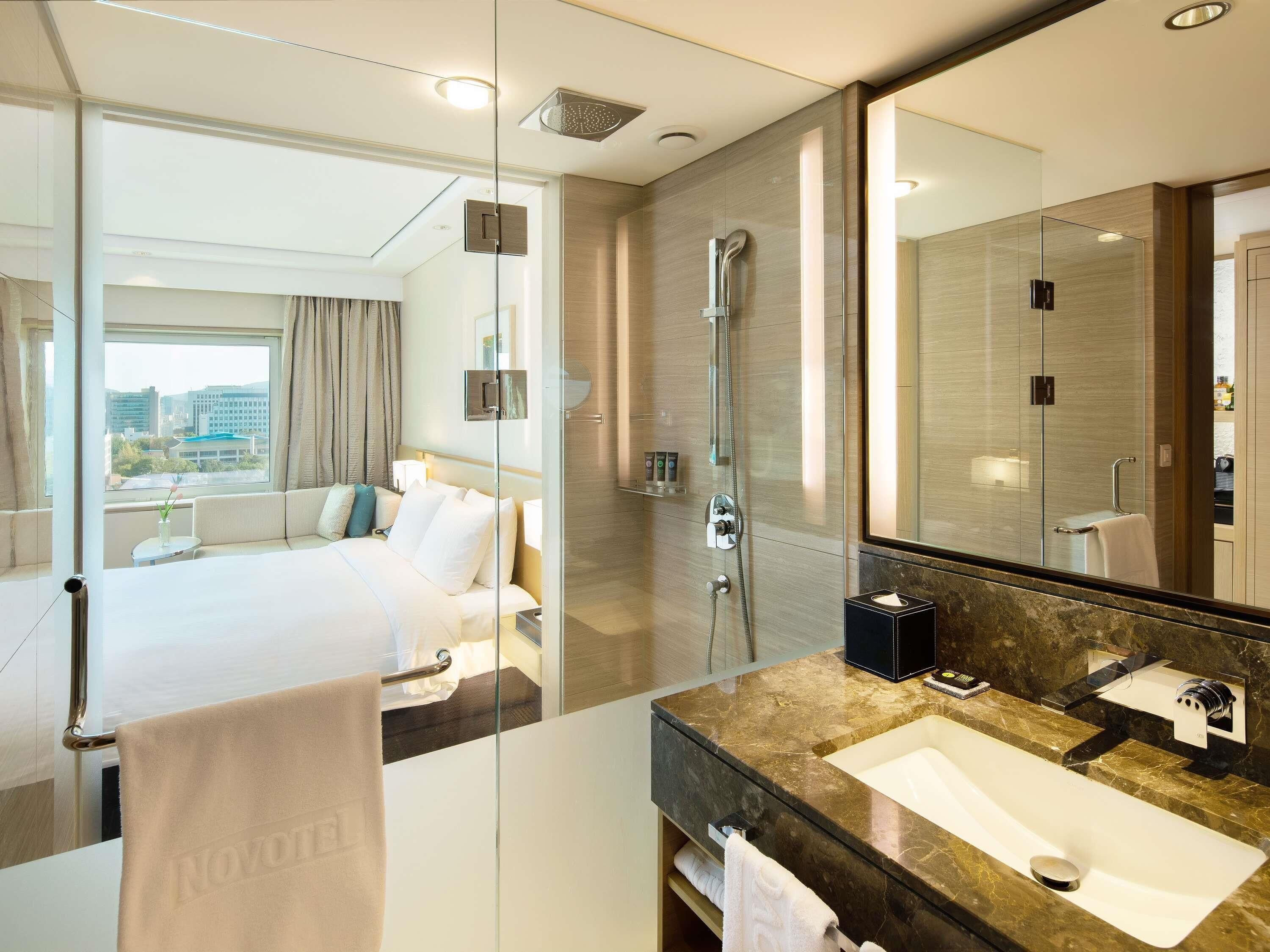 A hotel room with a bed, a shower, a large mirror, a sink, and a view of the city.