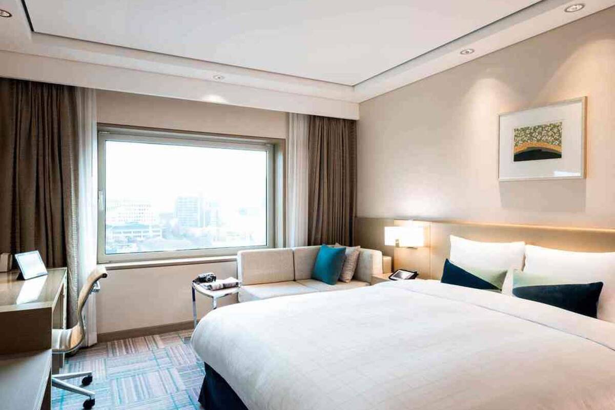 Une chambre d’hôtel avec un grand lit, un bureau, une chaise, une table de chevet, une lampe et une fenêtre avec des rideaux.