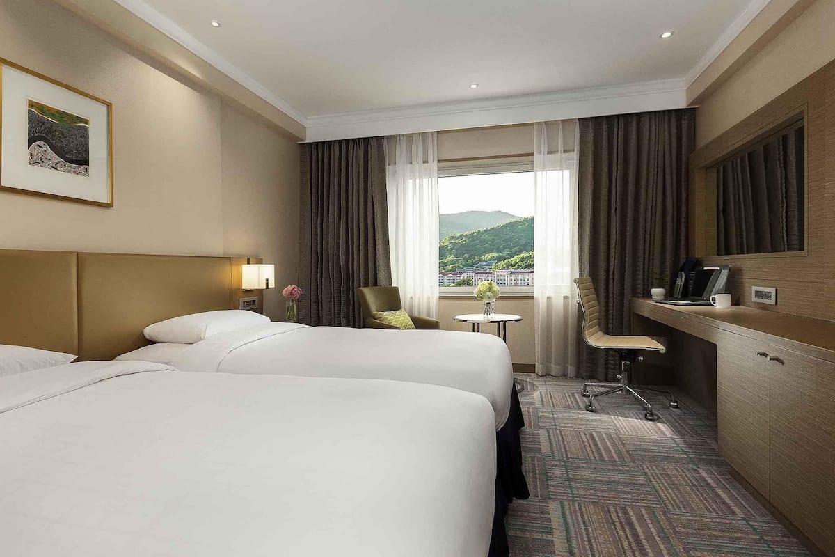 A hotel room with two beds, a desk, a chair, a small table with a vase, and a view of a mountain.