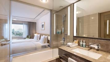 A modern hotel room with a glass-enclosed shower, a large bed, a separate bathroom with a marble sink, and a view of the city through the window.