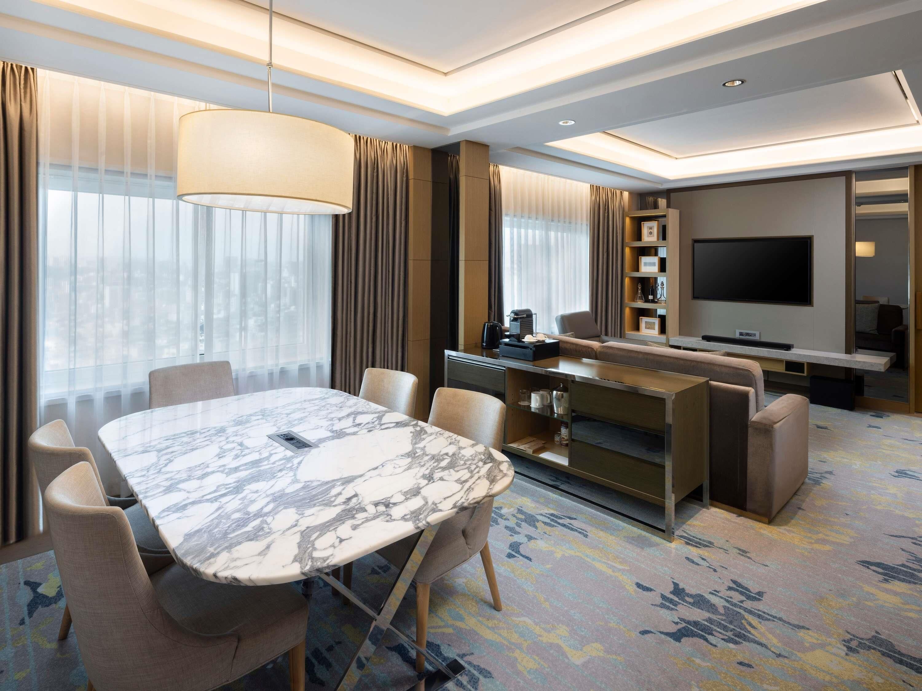 A modern hotel room with a marble dining table, comfortable seating, a sofa, a flat-screen TV, and a minibar.