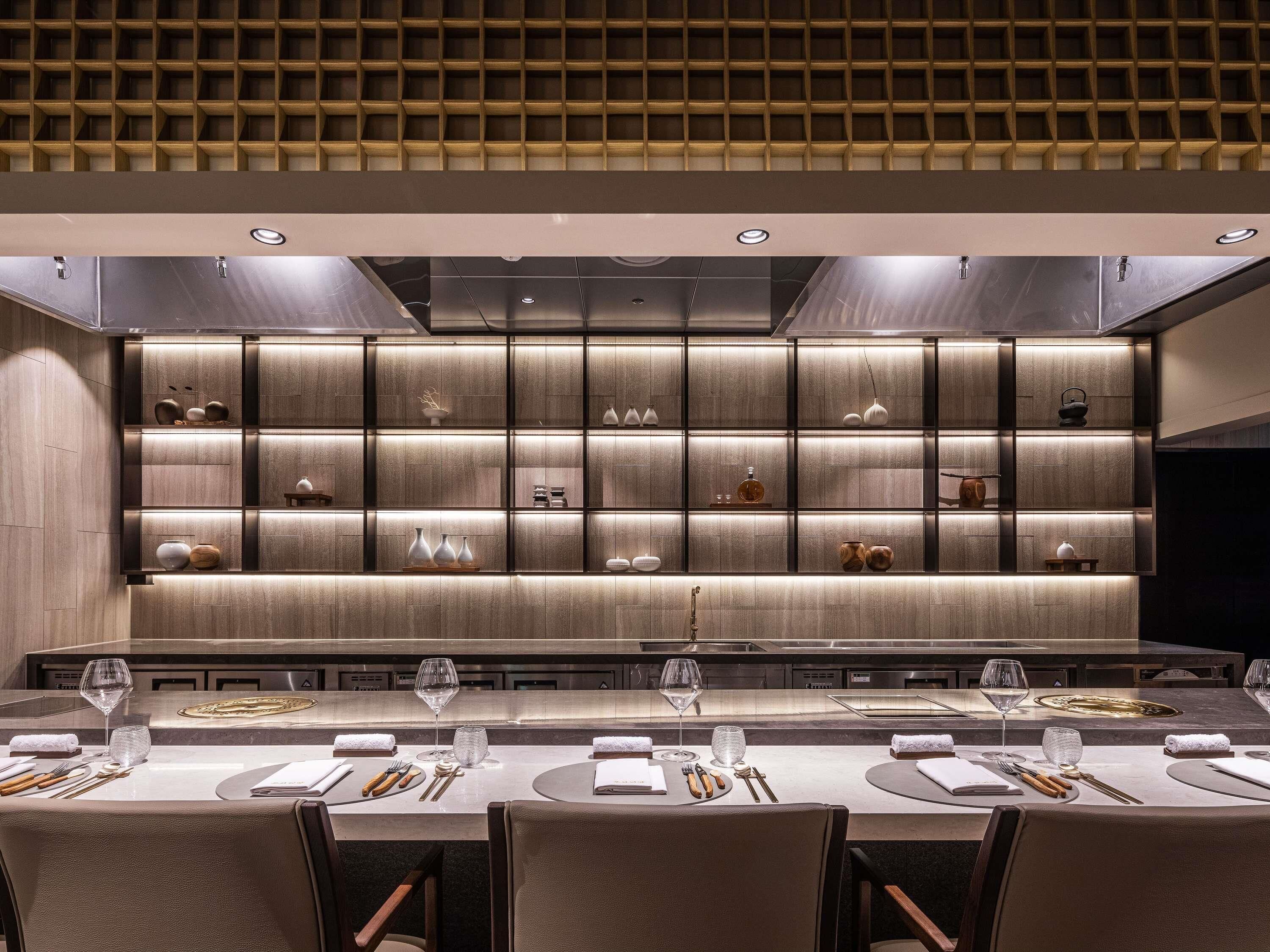 A modern dining area with a long table, multiple chairs, and a wall with shelves displaying various decorative items.