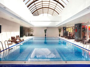 Una piscina interior con agua azul cristalina, rodeada de sillones de descanso y plantas en macetas.