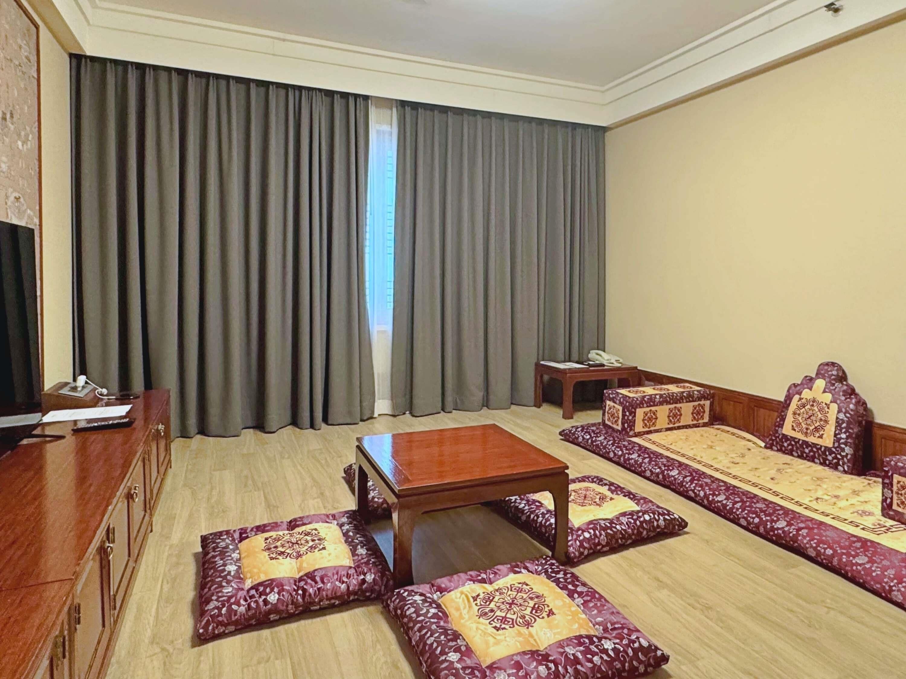 A hotel room with a large window, a sofa set, a wooden table, and a desk with a telephone.