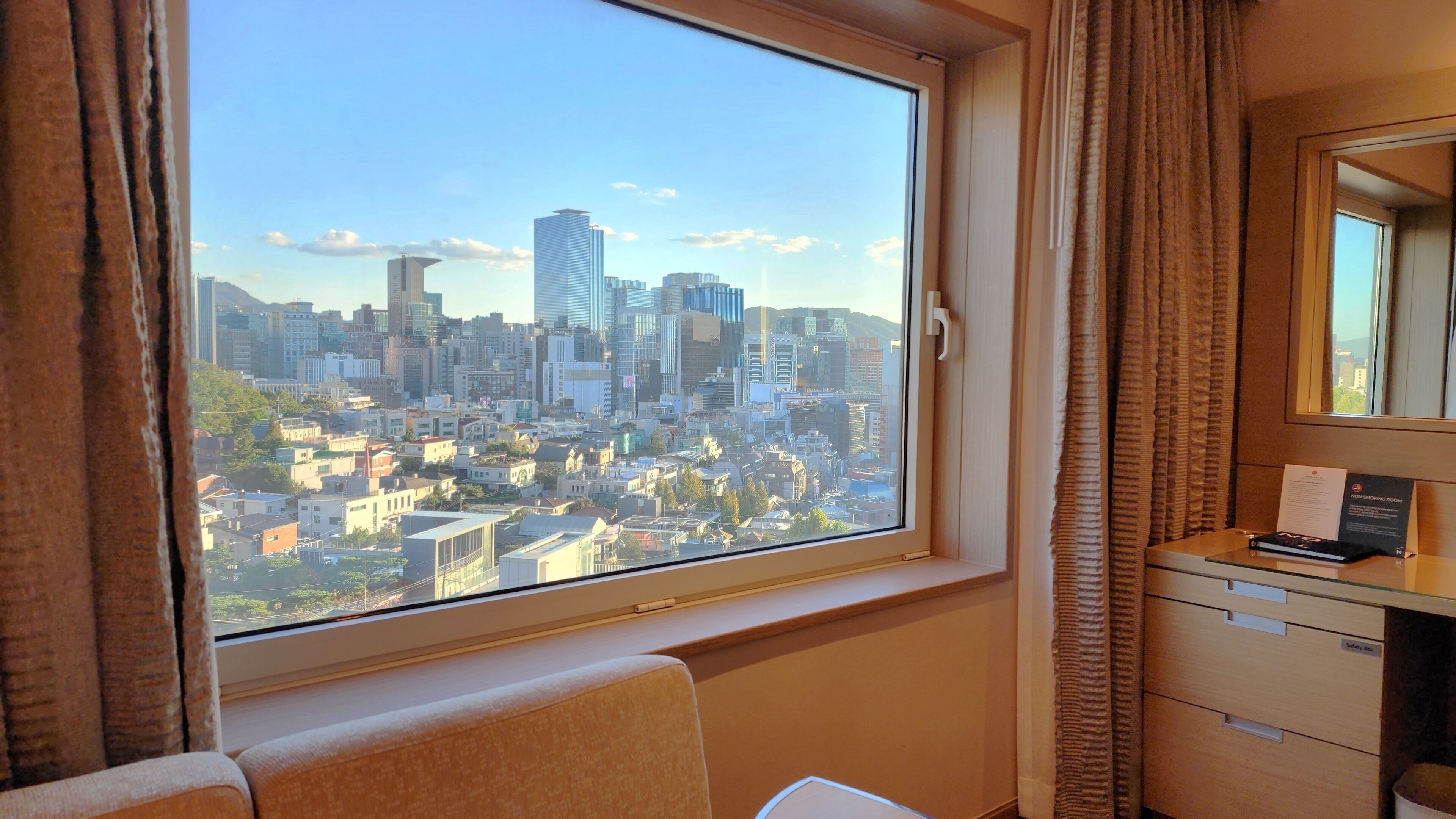 A hotel room with a large window offering a city view, a bedside table with a lamp, and a mirror on the wall.
