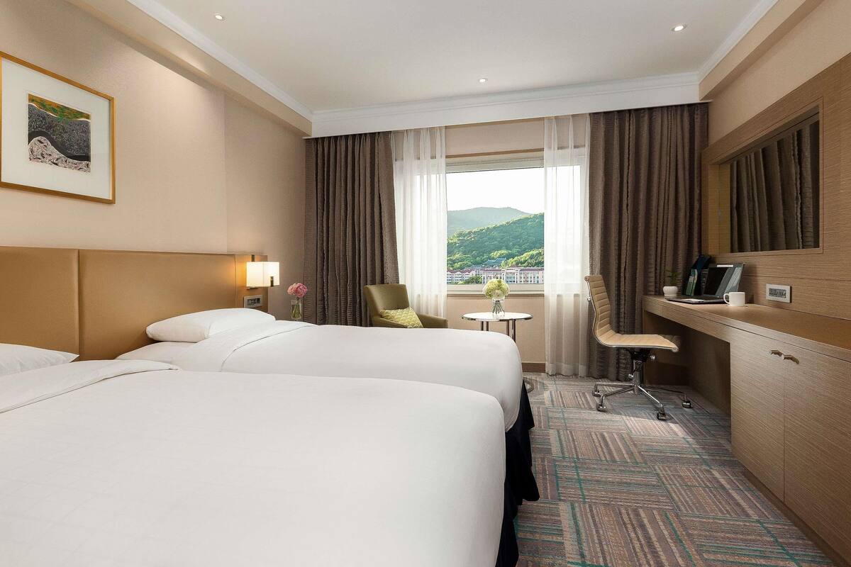 Une chambre d’hôtel avec deux lits, un bureau, une chaise, une petite table avec un vase et une fenêtre avec des rideaux.