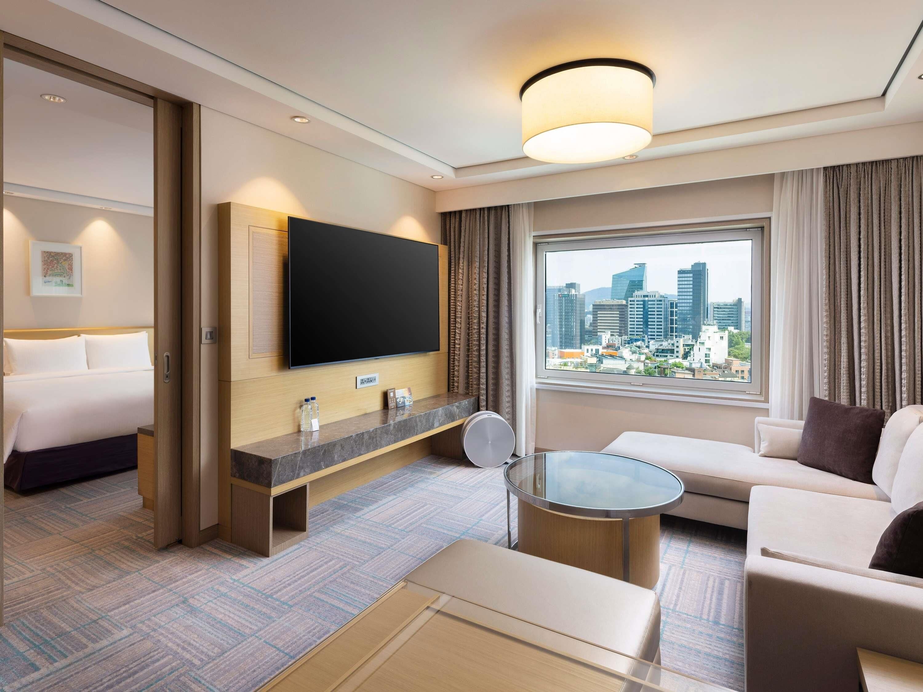A hotel room with a bed, a TV, a sofa, a coffee table, and a window with a city view.