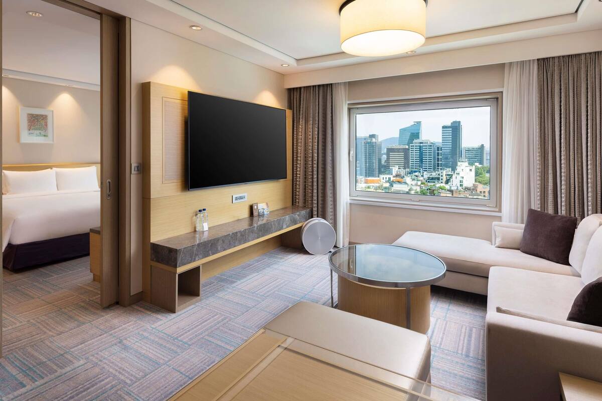 Une chambre d’hôtel équipée d’un lit, d’une télévision, d’un canapé, d’une table basse et d’une fenêtre donnant sur la ville.