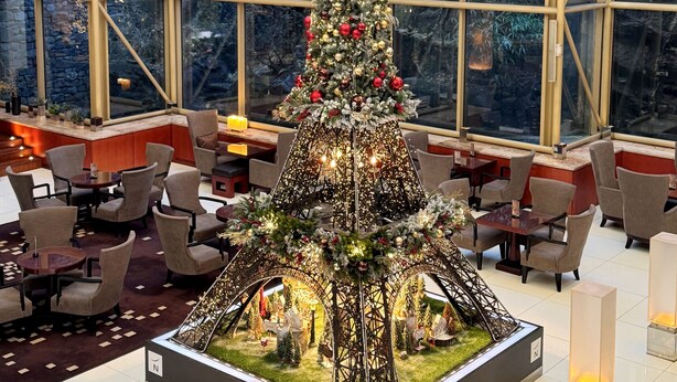 Een grote kerstboom in een ruimte, versierd met lichtjes en decoraties, omringd door zithoeken en een achtergrond van een stenen muur.